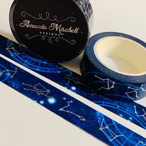 May include: Two rolls of decorative washi tape with a celestial theme. The tape features a dark blue background with silver constellations and a starry pattern. One roll displays the text "Amanda Mischell DESIGNS".