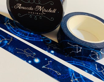 Puede incluir: Dos rollos de cinta adhesiva decorativa con temática celestial. La cinta presenta un fondo azul oscuro con constelaciones plateadas y un patrón de estrellas. Un rollo muestra el texto "Amanda Mischell DESIGNS".