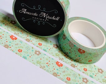 Puede incluir: Dos rollos de cinta washi con un patrón floral. La cinta es verde con flores naranjas y marrones. La cinta está sobre un fondo blanco. La cinta está etiquetada "Amanda Mischell Designs".