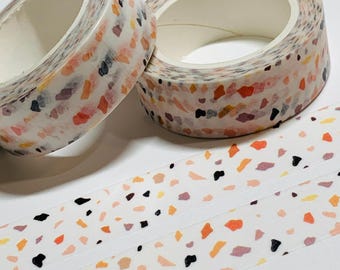 Puede incluir: Dos rollos de cinta adhesiva decorativa con un patrón de terrazo en naranja, negro y morado sobre un fondo blanco. Una tira de la misma cinta está desenrollada debajo, mostrando el patrón. La cinta es ideal para manualidades y scrapbooking.