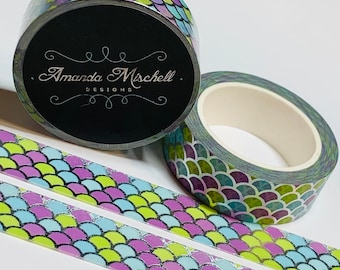 May include: Two rolls of washi tape with a mermaid scale pattern in shades of green, purple, and blue. The tape is on a white surface. The label on the roll reads "Amanda Mischell Designs."