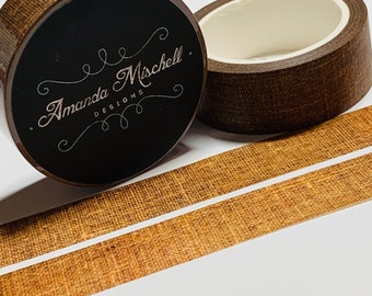 May include: Two rolls of brown washi tape with a burlap texture. The tape is on a white surface. One roll has a black label with the text "Amanda Mischell Designs".