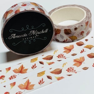 May include: Two rolls of washi tape with a white background and a pattern of autumn leaves in shades of brown, orange, and yellow. The tape is labeled "Amanda Mischell Designs".