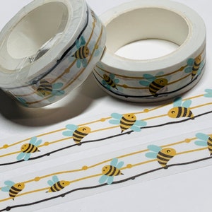 May include: Two rolls of decorative washi tape with a repeating pattern of cartoon bees. The bees are yellow and black with light blue wings, set against a white background with black and gold lines. The tape is ideal for crafts and scrapbooking.