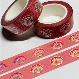 May include: Two rolls of red washi tape with a seashell pattern, along with two strips of the tape. The tape features a repeating design of white and gold seashells. The background of the tape is a solid red color.