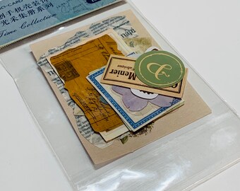 May include: A clear plastic package containing a collection of decorative paper ephemera. The ephemera includes vintage-style labels, tags, and paper scraps with text and floral designs. The package also features text in multiple languages, including "Time Collection."