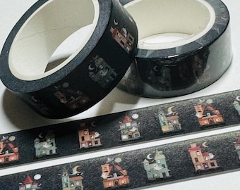 May include: Two rolls of black and white washi tape with a repeating pattern of spooky houses and crescent moons. The tape is on a black surface.