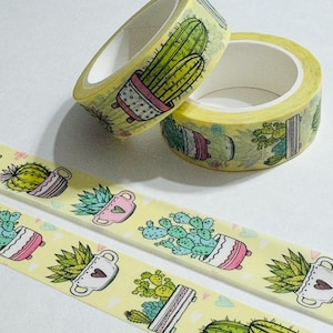 May include: Two rolls of yellow washi tape with a cactus design. The tape features illustrations of cacti in pots, with pink, white, and green accents. Two strips of the same tape are visible below the rolls.