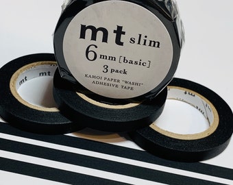 Puede incluir: Rollos de cinta adhesiva washi negra mt slim, de 6 mm de ancho, en un paquete de 3. El embalaje incluye el texto "KAMOI PAPER WASI ADHESIVE TAPE". Varios rollos son visibles, con cinta desenrollada en rayas blancas y negras.