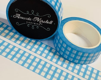May include: Two rolls of blue and white gingham patterned washi tape. One roll is upright, displaying the black label with the text "Amanda Mischell Designs". The other roll is on its side. Two strips of the tape are unrolled.