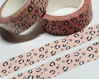 Puede incluir: Dos rollos de cinta adhesiva decorativa con un diseño de estampado de leopardo en tonos marrón y negro. Dos tiras de la misma cinta están desenrolladas, mostrando el patrón repetido. La cinta está sobre una superficie blanca.