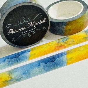 May include: Two rolls of adhesive tape featuring a watercolor pattern of blended blue and yellow hues. One roll displays a black circular label with the white text Amanda Mischell DESIGNS surrounded by decorative white flourishes.