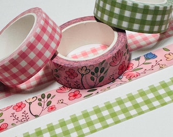 May include: Three rolls of decorative washi tape in various designs. One roll features a pink and white gingham pattern, another has a floral design with pink flowers, and the third has a green and white gingham pattern. Two strips of tape are also visible.