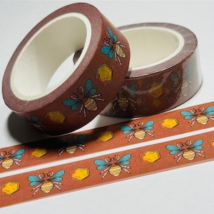 May include: Two rolls of decorative washi tape with a brown background. The tape features a repeating pattern of bees with blue wings and yellow honeycomb shapes. A few strips of the tape are also visible.