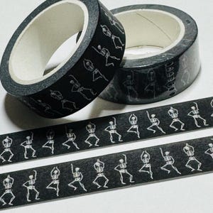 May include: Two rolls of black washi tape with a white skeleton design. The skeletons are in a dancing pose.
