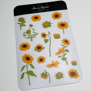 May include: A sheet of stickers featuring various illustrations of sunflowers in different poses and sizes. The stickers are white with a light gray outline. The sheet is labeled "Amanda Mitchell Designs."