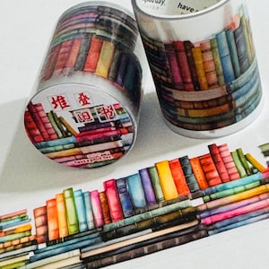 May include: Two rolls of decorative washi tape featuring a colorful watercolor design of stacked books. The tape rolls are in clear plastic containers, with text in a foreign language and the phrase "have a good day". A strip of the tape is unrolled.