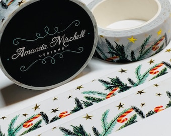 Può includere: Un rotolo di nastro washi bianco con un design in foglia d'oro che presenta rami di pino verdi, bastoncini di zucchero rosso e bianco e stelle dorate. Il nastro è etichettato "Amanda Mischell Designs".