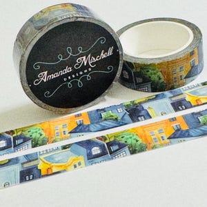 May include: A roll of decorative washi tape featuring a watercolor illustration of colorful townhouses and green trees. The black circular label on the side of the roll displays the white text Amanda Mischell DESIGNS with decorative flourishes.