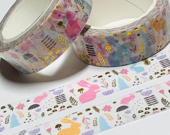 Puede incluir: Dos rollos de cinta washi con fondo blanco y un patrón repetido de conejos rosas y amarillos, setas doradas y diseños florales. La cinta está decorada con detalles de lámina dorada.