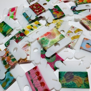 May include: A collection of colorful washi tapes with various fruit and floral designs, displayed on white plastic spools. The tapes are arranged in a random pattern, showcasing a variety of designs.