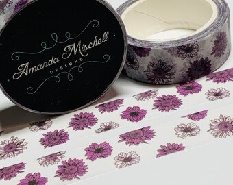 May include: A roll of washi tape with a floral pattern. The tape is white with purple flowers and a light gray background. The tape is on a white surface. The tape is labeled "Amanda Mischell Designs".