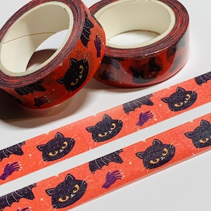 May include: Two rolls of Halloween-themed washi tape with a repeating pattern of black cats, bats, and purple hands on an orange background. The tape is designed for crafting and decorative purposes.
