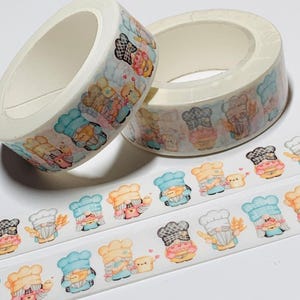May include: Two rolls of decorative washi tape featuring a repeating pattern of cartoon chef gnomes. The gnomes wear chef hats in various colors and hold baked goods. The tape is white with a light blue border.