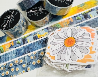 May include: Three rolls of decorative washi tape with black labels reading Amanda Michell Designs, alongside strips of printed tape featuring denim, daisy, and building patterns, and a clear plastic bag containing daisy-shaped stickers.