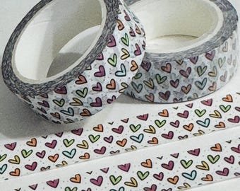 Puede incluir: Dos rollos de cinta adhesiva decorativa con fondo blanco y un patrón de corazones coloridos. La cinta tiene un borde gris y está parcialmente desenrollada, revelando el patrón de corazones. También se ven dos tiras de cinta.