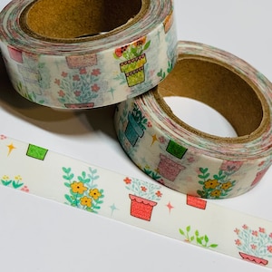 May include: Two rolls of washi tape with a floral pattern featuring potted plants and flowers. The tape is white with a light pink, green, and yellow color scheme.