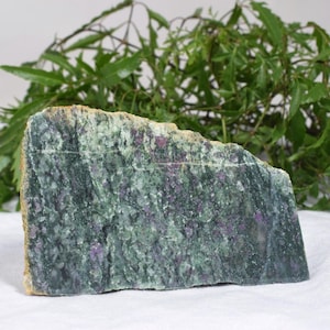 May include: A flat, rectangular slab of zoisite mineral featuring a mottled green base with embedded, irregular patches of deep magenta-colored ruby crystals. The stone has a rough, natural texture with visible mineral inclusions throughout the surface.