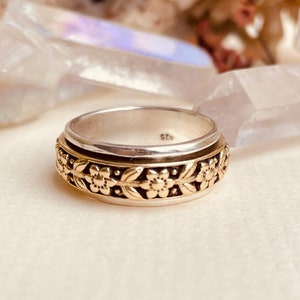 Könnte beinhalten: Ein silberner und goldener Spinnerring mit floralem Design. Der Ring besteht aus zwei Bändern, einem silbernen und einem goldenen, die sich unabhängig voneinander drehen. Das florale Design befindet sich auf dem goldenen Band.