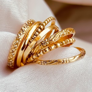 May include: A collection of gold rings with various textures and designs, including beaded, floral, and engraved patterns. The rings are stacked together on a white satin surface, showcasing their intricate details and warm golden hue.