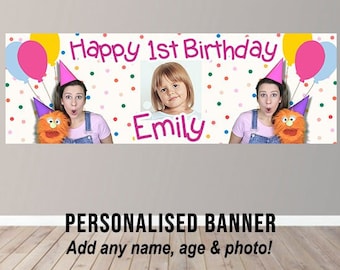 May include: Personalized birthday banner with a colorful confetti background. The banner says "Happy 1st Birthday Emily" and features a photo of a young girl. There are two women holding puppets on either side of the banner.