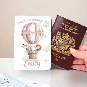 May include: A personalised passport holder with a pink hot air balloon design, the text "The Adventures of Emily", and a passport. The passport holder features a giraffe, elephant, and lion in a basket. The passport is burgundy.