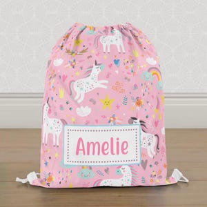 May include: Pink drawstring bag patterned with white unicorns, rainbows, stars, and flowers. The bag has the name "Amelie" in pink on a white label. The bag is made of a soft fabric. The bag is suitable for children.