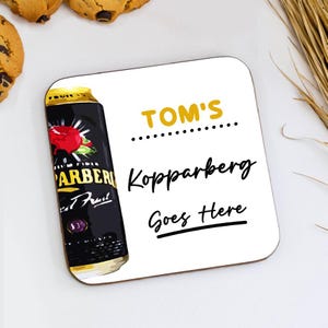 May include: A square white coaster with rounded corners features a black and gold can of Kopparberg cider. The coaster has the text "TOM'S", "Kopparberg", and "Goes Here" in black and gold.