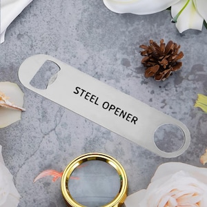 Op de afbeelding: Een roestvrijstalen flesopener met de zwarte tekst "STEEL OPENER". De opener ligt op een grijze ondergrond, met een vergrootglas met gouden rand en een dennenappel op de achtergrond.