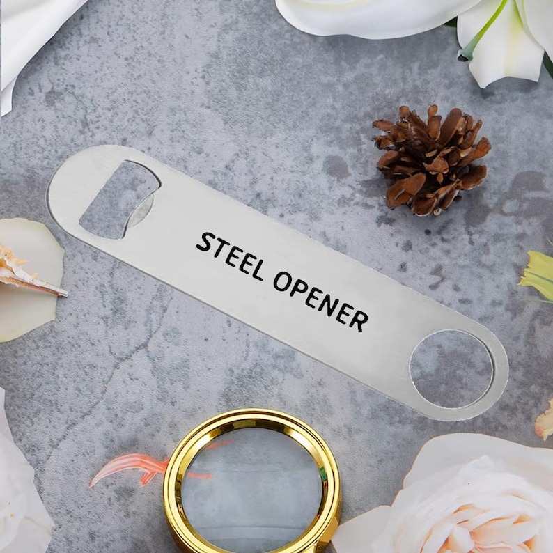 Op de afbeelding: Een roestvrijstalen flesopener met de zwarte tekst "STEEL OPENER". De opener ligt op een grijze ondergrond, met een vergrootglas met gouden rand en een dennenappel op de achtergrond.