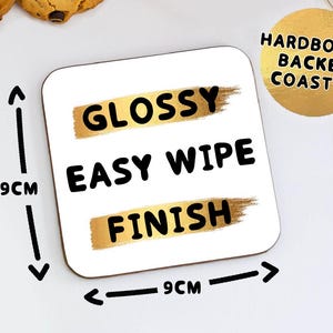 May include: Square white coaster with the words "GLOSSY EASY WIPE FINISH" in black text. The coaster is 9cm x 9cm and has a gold-colored brushstroke design. The coaster is labeled "HARDBOARD BACKED COASTER".