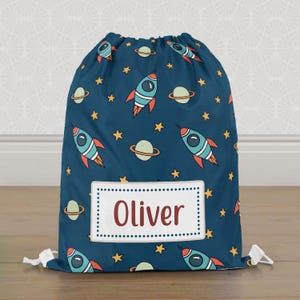 May include: A navy blue drawstring bag with a space-themed design of rockets, planets, and stars. The bag has a white label with the name "Oliver" in red. White drawstrings are visible at the top, and the bag is on a wooden floor.