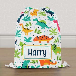 May include: A white drawstring bag with a vibrant dinosaur print. The bag includes a rectangular label with the name "Harry" in navy blue. The dinosaurs are in various colors, including green, orange, yellow, and pink.