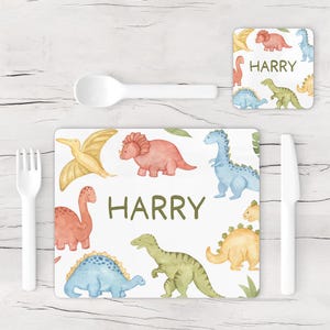 May include: A white placemat, coaster, spoon, fork, and knife set. The placemat and coaster feature colorful watercolor dinosaurs and the name "HARRY" in green. The dinosaurs are in shades of red, blue, yellow, and green.