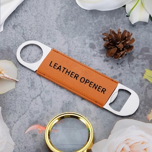 Op de afbeelding: Een bruine leren flesopener met de tekst "LEATHER OPENER" in het zwart. De opener heeft een roestvrijstalen flesopener en een ring om op te hangen. De achtergrond is een grijs oppervlak met bloemaccenten.
