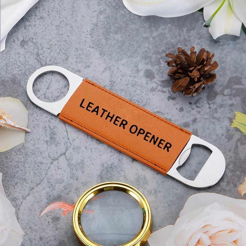 Op de afbeelding: Een bruine leren flesopener met de tekst "LEATHER OPENER" in het zwart. De opener heeft een roestvrijstalen flesopener en een ring om op te hangen. De achtergrond is een grijs oppervlak met bloemaccenten.