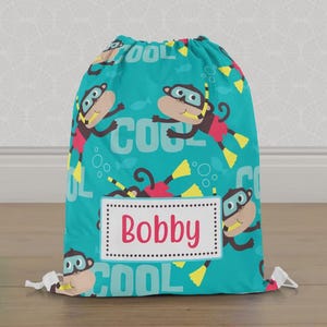 May include: A turquoise drawstring bag with a pattern of cartoon monkeys wearing snorkels and flippers. The word "COOL" is printed in white. A rectangular label with the name "Bobby" in red is centered on the bag.