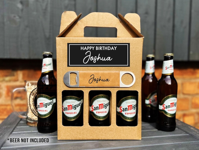 Op de afbeelding: Een bruine kartonnen bierdrager met een handvat, met drie flessen San Miguel-bier. De drager heeft een zwart label met de tekst "Happy Birthday Joshua". Een flesopener is inbegrepen. De tekst "*BEER NOT INCLUDED" staat onderaan.