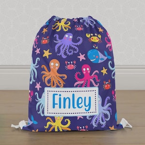 May include: A navy blue drawstring bag with a vibrant sea creature design. The pattern includes octopuses, crabs, starfish, and a whale. The bag has the name "Finley" in a white rectangle with a blue border.