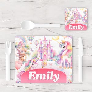May include: A children's placemat and coaster set with a unicorn theme. The placemat shows a castle, rainbows, and unicorns, with the name "Emily" in a pink banner. A white spoon, fork, and knife are included.
